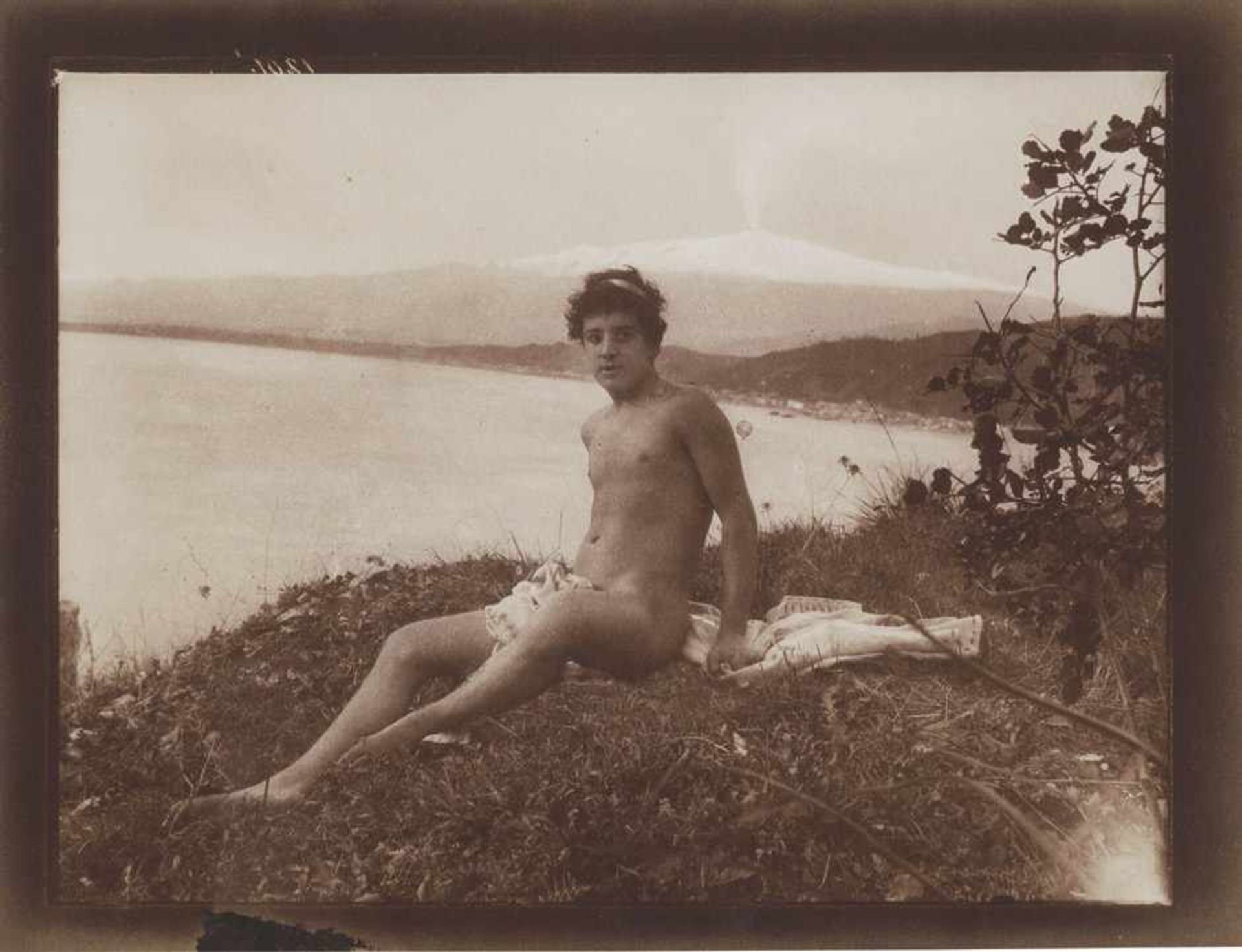 Gloeden, Wilhelm von: Young nude male sitting on grass, Etna in the background Young nude boy