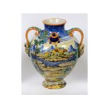 A maiolica drug jar, decorated figures in a landscape, with two spouts and faun mask handles, chips,