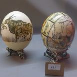 Two ostrich eggs one hand painted with buffalo the other printed with elephants both on white metal