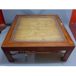 A contemporary large teak coffee table with rattan top under glass raised on block legs joined by