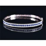 A white metal, diamond, and sapphire bangle channel-set with a line of round brilliant cut sapphires