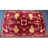 A large wool floor rug, decorated with bridles, rosettes and ribbons in gold on a dark red ground,