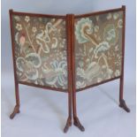 An Edwardian mahogany firescreen, inset with two crewel work and glazed panels, on end supports,