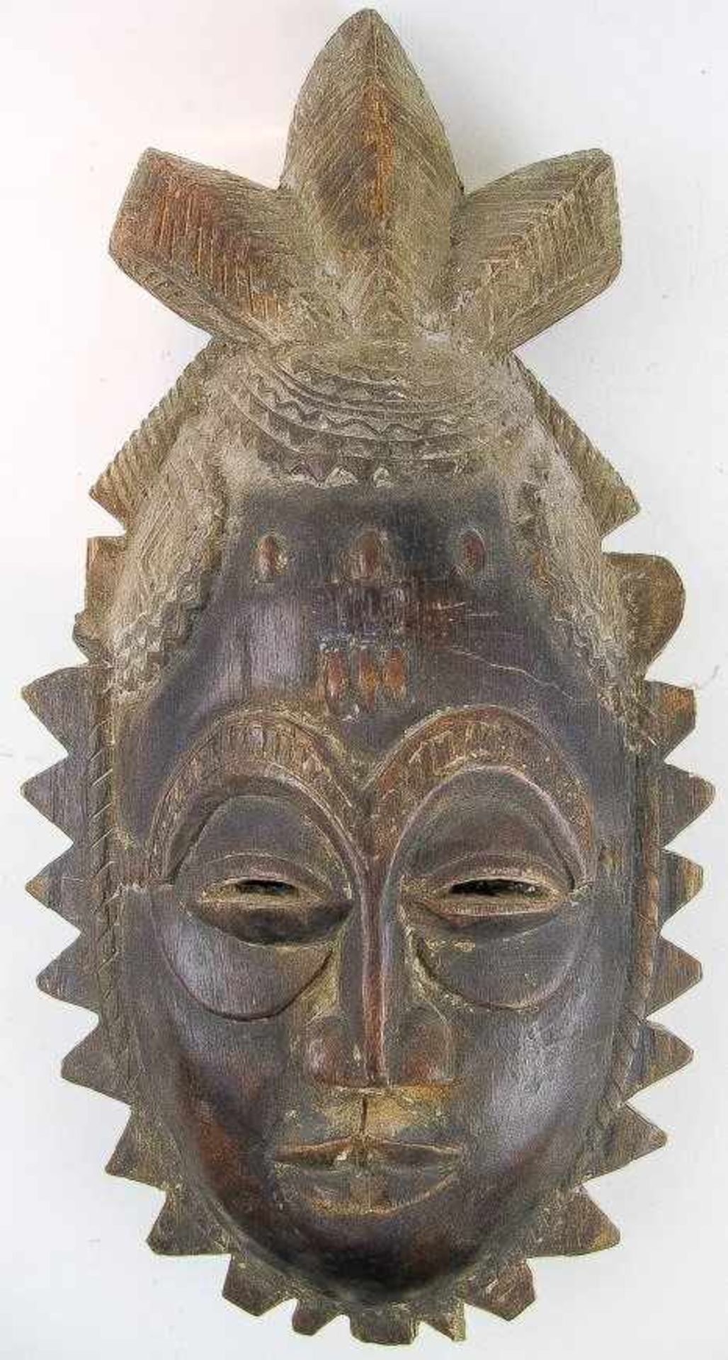 Maske. Holz. Menschliches Gesicht in zackenförmiger Umrahmung. Frisur aus drei großen Knoten. H.