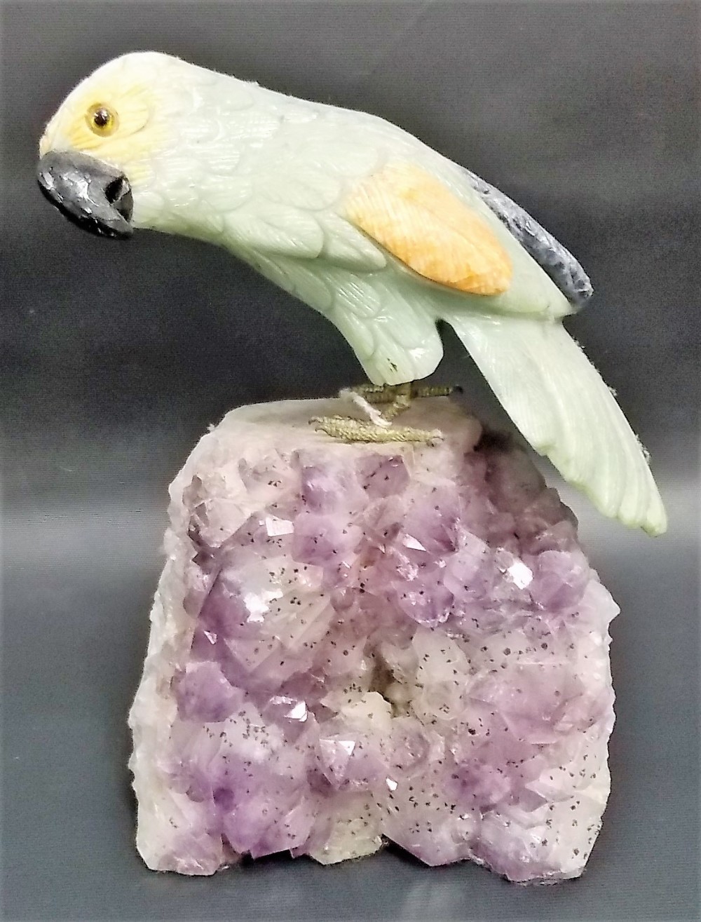 Interesting hard stone model of a parrot with glass inset eyes upon an amethyst specimen boulder,