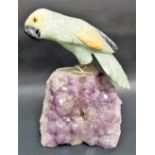 Interesting hard stone model of a parrot with glass inset eyes upon an amethyst specimen boulder,