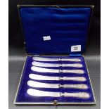 Cased set of six Edwardian silver embossed weighted handle butter knives, the handles foliate