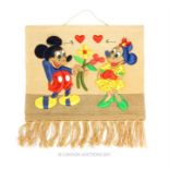 A wall hanging, decorated with Mickey Mouse handing Minnie Mouse a bouquet of flowers