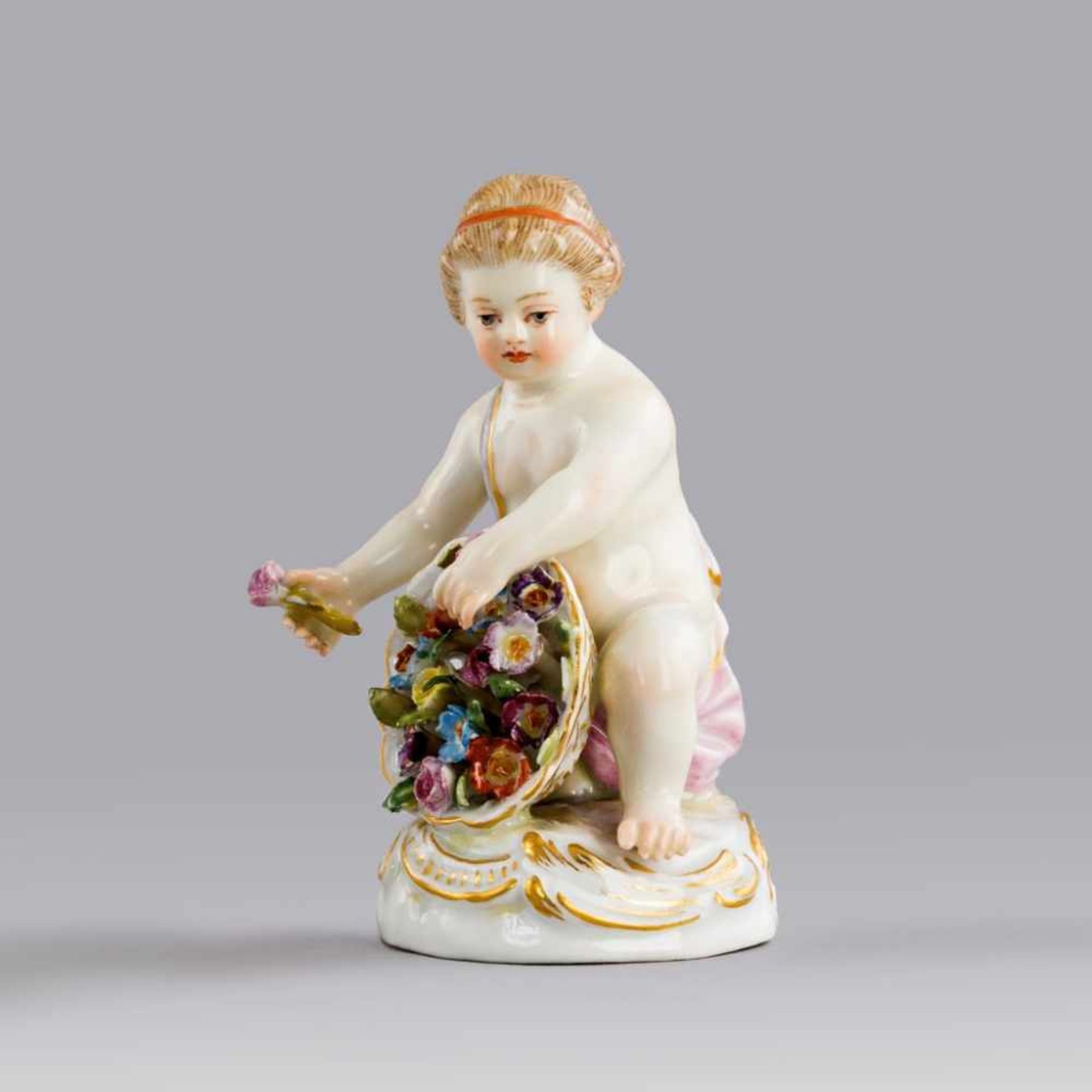Kniende Amorette mit Blütenkorb. Polychrom staffierte Figurine auf Rocaillensockel. Blaue