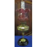 Brass oil lamp with pedestal support on ceramic base, cranberry coloured shade with clear chimney.