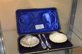 A pair of Edwardian silver shell form butter dishes, Birmingham 1903 and 1904, cased with knives (