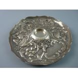 A CIRCULAR SILVER INK BOTTLE TRAY, heavily scrolled with raised flowers and leaves and with wavy