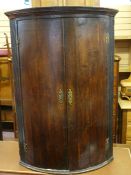 A GEORGIAN OAK BOW FRONT HANGING CORNER CUPBOARD, the doors with brass 'H' hinges and decorative