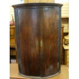 A GEORGIAN OAK BOW FRONT HANGING CORNER CUPBOARD, the doors with brass 'H' hinges and decorative