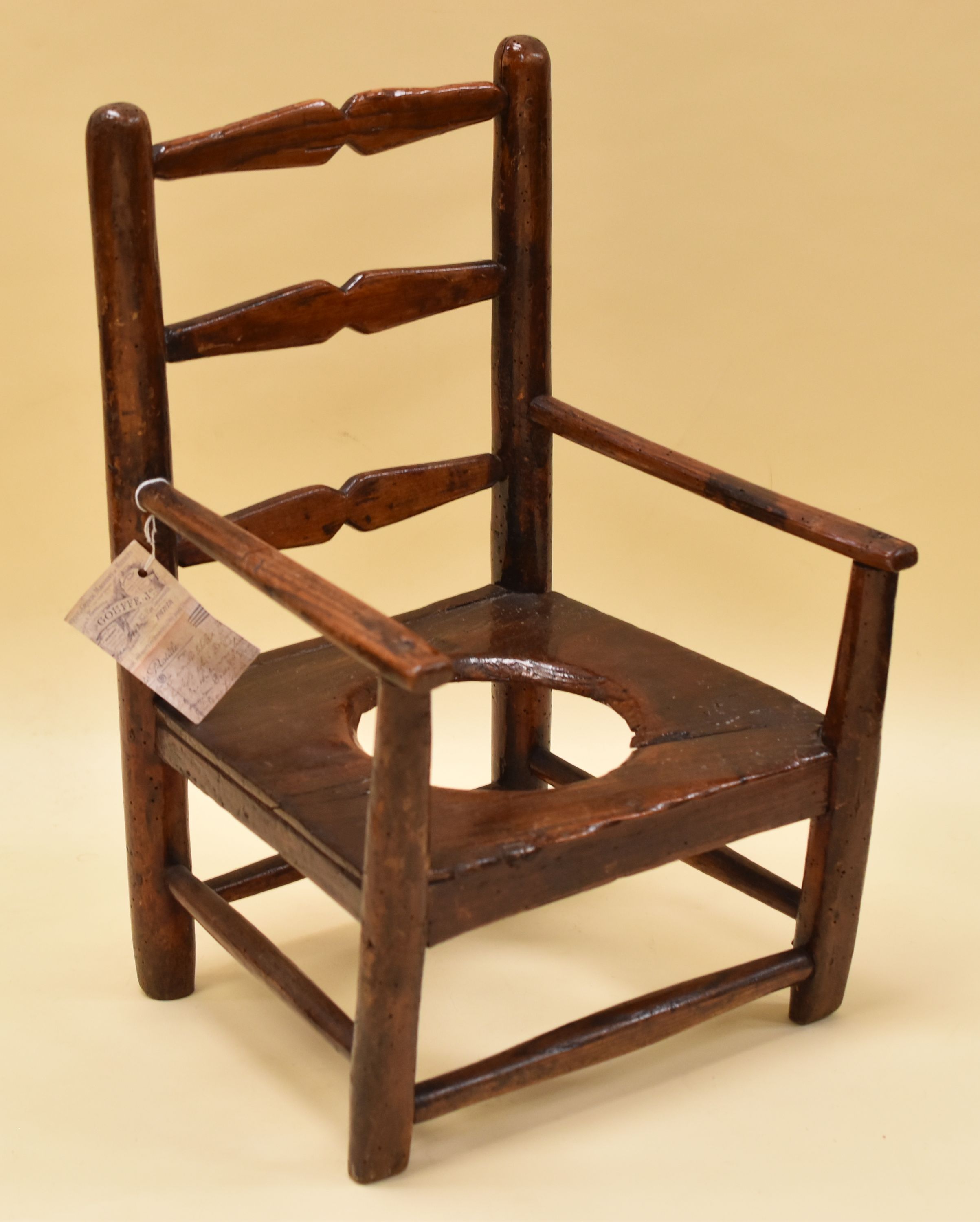 A INFANT'S PRIMITIVE POTTY CHAIR possibly in elm, with three rung ladder back and arm supports,