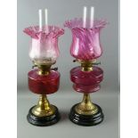 TWO VICTORIAN OIL LAMPS, both brass columned with cranberry glass shades and reservoirs, one