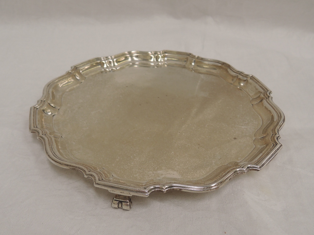 A silver waiter of plain form having a raised shaped pie crust rim and trefoil scroll feet,