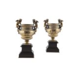 A PAIR OF BRONZE URNS with flanking putto and basket weave banding on square marble bases. 32cm