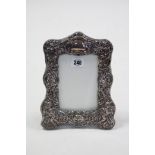 A silver rectangular photograph frame with embossed cherub, mask, & scroll design & on easel