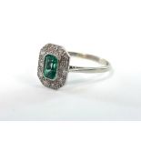 A platinum ring set rectangular emerald within a border of ten small diamonds.