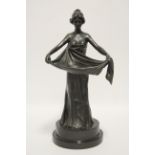 A bronze standing figure of a young lady wearing long flowing dress & holding the folds in front