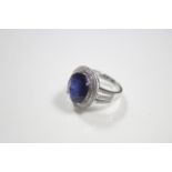 A white 14K ring set large blue oval-cut gemstone within a double border of small diamonds.