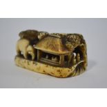 A stained ivory netsuke, carved as an elephant beside figures and two thatched dwellings,