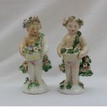 A pair of Derby cherub figurines holding a basket of flowers with flowers and leaves around,