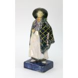 A ceramic figure of a woman wearing a cape and a wide brimmed hat on a rectangular blue plinth base,