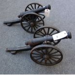 A pair of modern cast iron cannons, each circular tapering barrel on a carriage and spoked wheels,