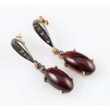 A pair of garnet and diamond drop stud earrings, each set with an oval garnet cabochon, surmounted