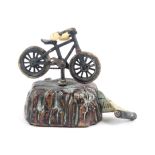 A patinated brass novelty tape measure as a bicycle on rocky outcrop, complete printed tape in ins