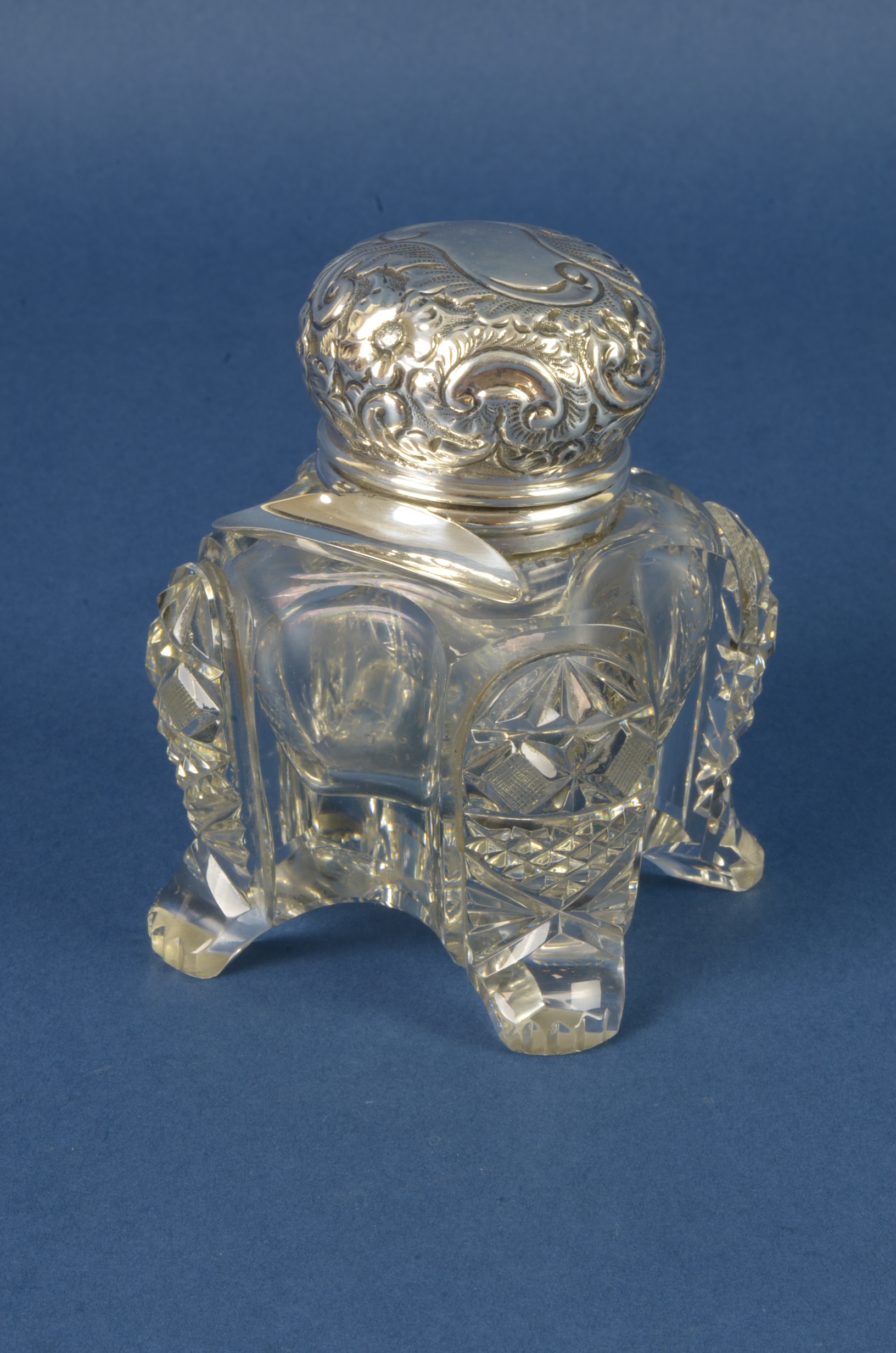 A cut glass Inkwell with floral and scroll embossed hinge lid and pen shelf, mounted upon four paw