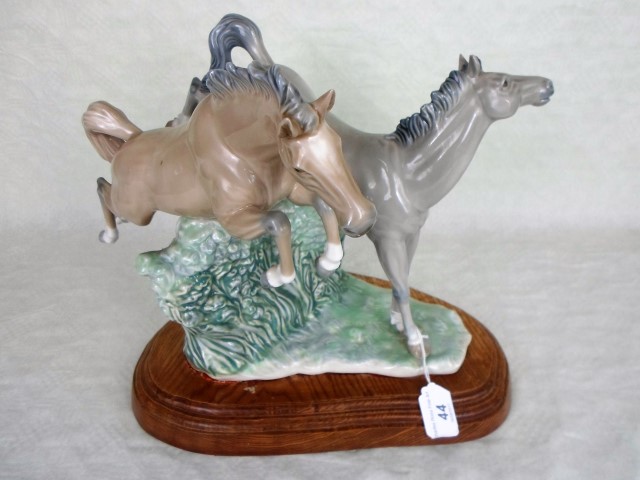 A Nao figure group of two horses jumping a hedge, mounted to bespoke wooden stand, 29cm tall.