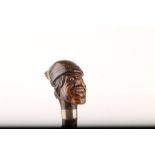 AN INTERESTING FOLK ART WOOD CANE, modelled as a man's head wearing a working hat, early glass eyes,