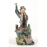 A CHINESE PORCELAIN FIGURE OF PINDOLA. Late Qing Dynasty. Seated against a rocky outcrop, with the