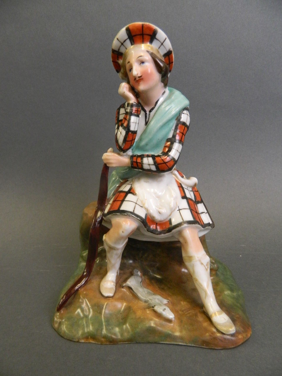 A porcelain figure of a Scottish highlander seated on a rock with a small dog asleep at his feet,