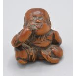 A Japanese wooden netsuke of a seated boy holding a mask behind his back and making a bekkako
