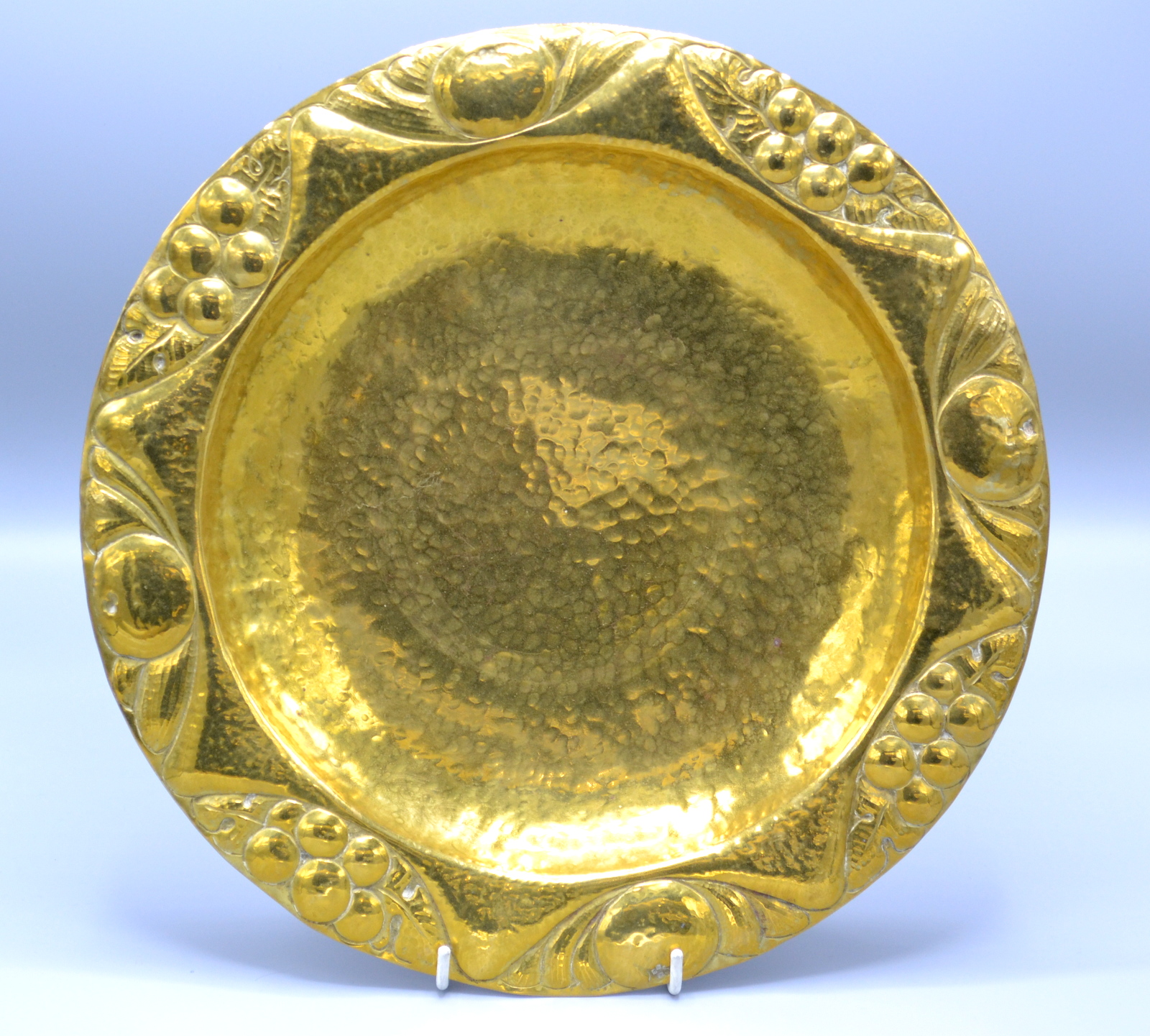 A brass Arts and Crafts dish with a fruit decorated repousse rim and planished centre, impressed F.