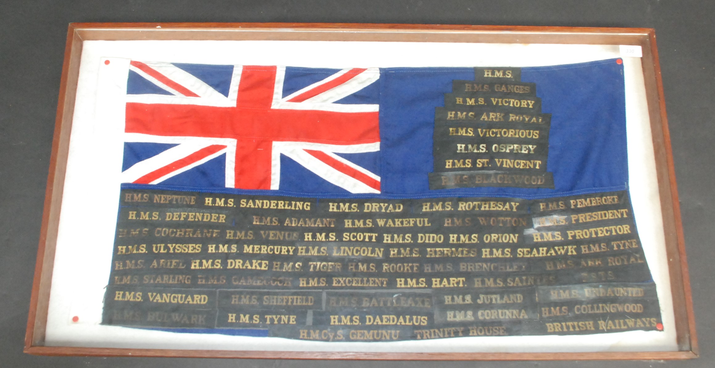 A blue ensign flag decorated with naval cap tallies, all in a mahogany framed display case,