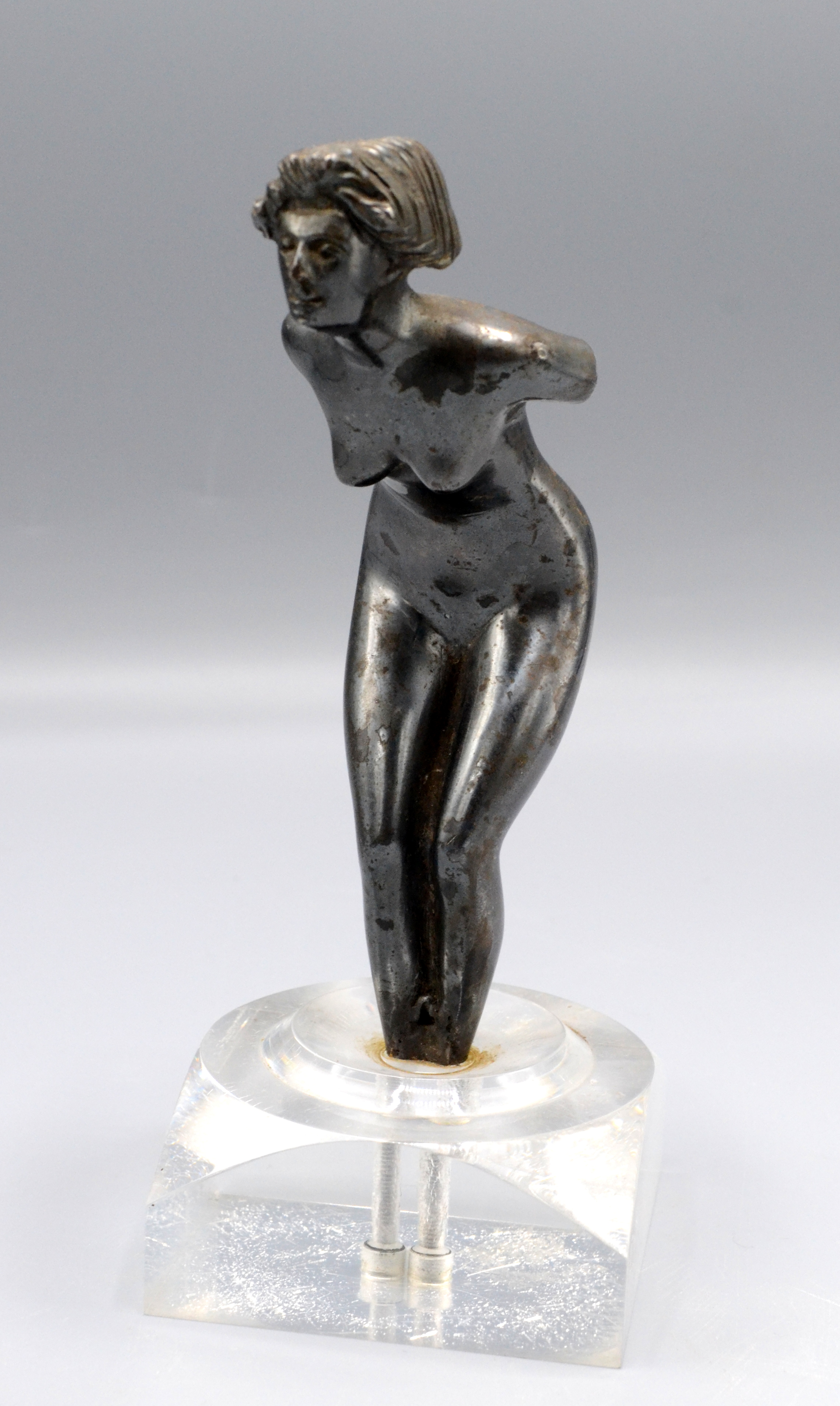 A white metal car mascot in the form of a naked lady mounted on a perspex base, height 16cm.