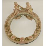 A German porcelain oval wall mirror, surmounted by two winged cherubs above a rose decorated frame,