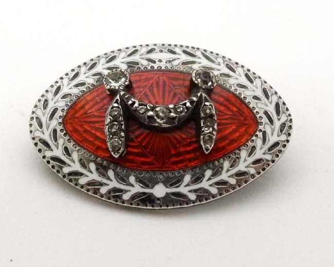 A white metal brooch of elliptical form with white and red gullioche enamel decoration and paste