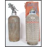 Two vintage oversized Soda Syhons by Sparklets, both with wire mesh covers and one retaining its