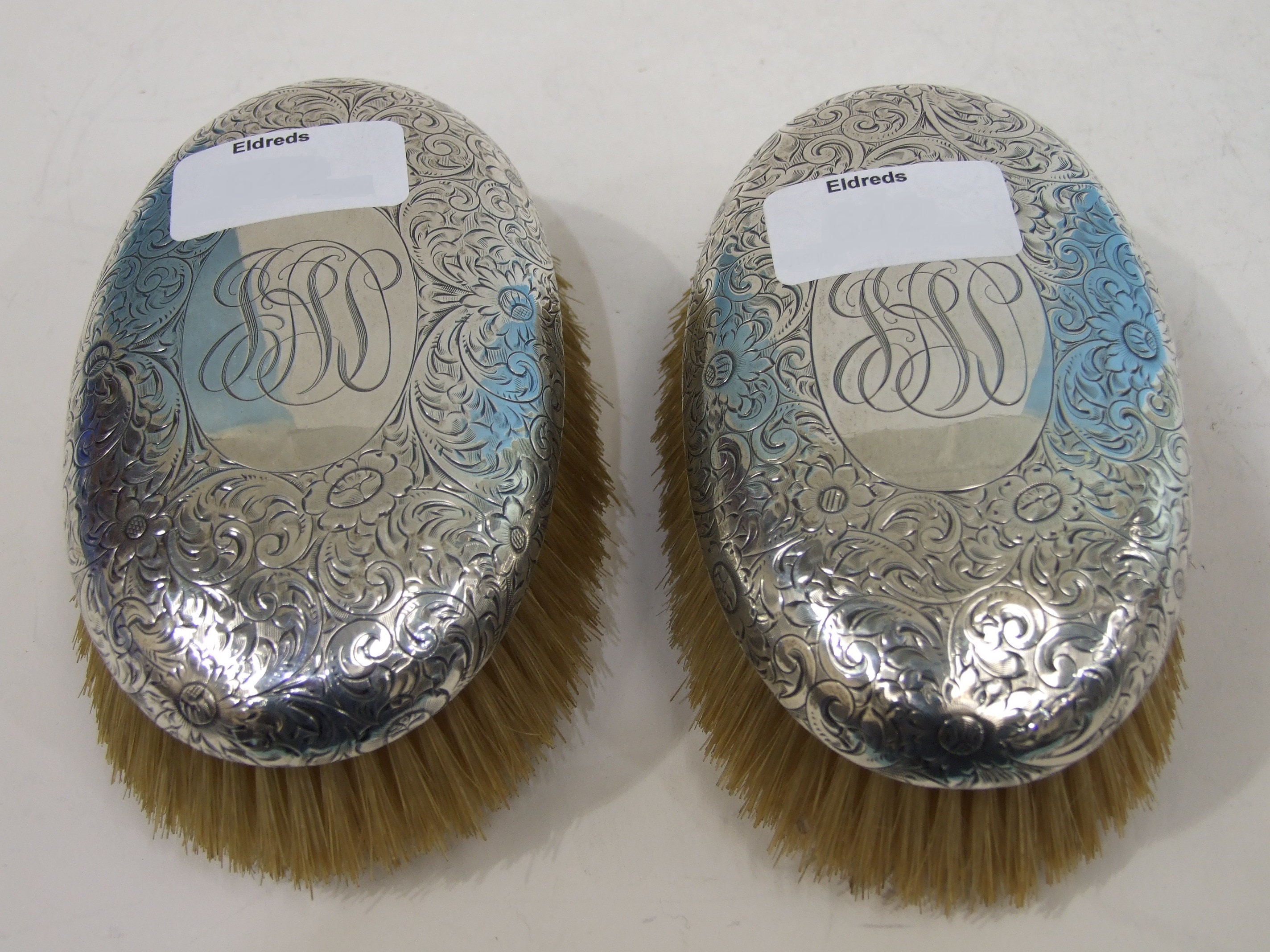 A pair of white-metal-backed oval hair brushes with overall foliate engraving, 14cm long, (2).