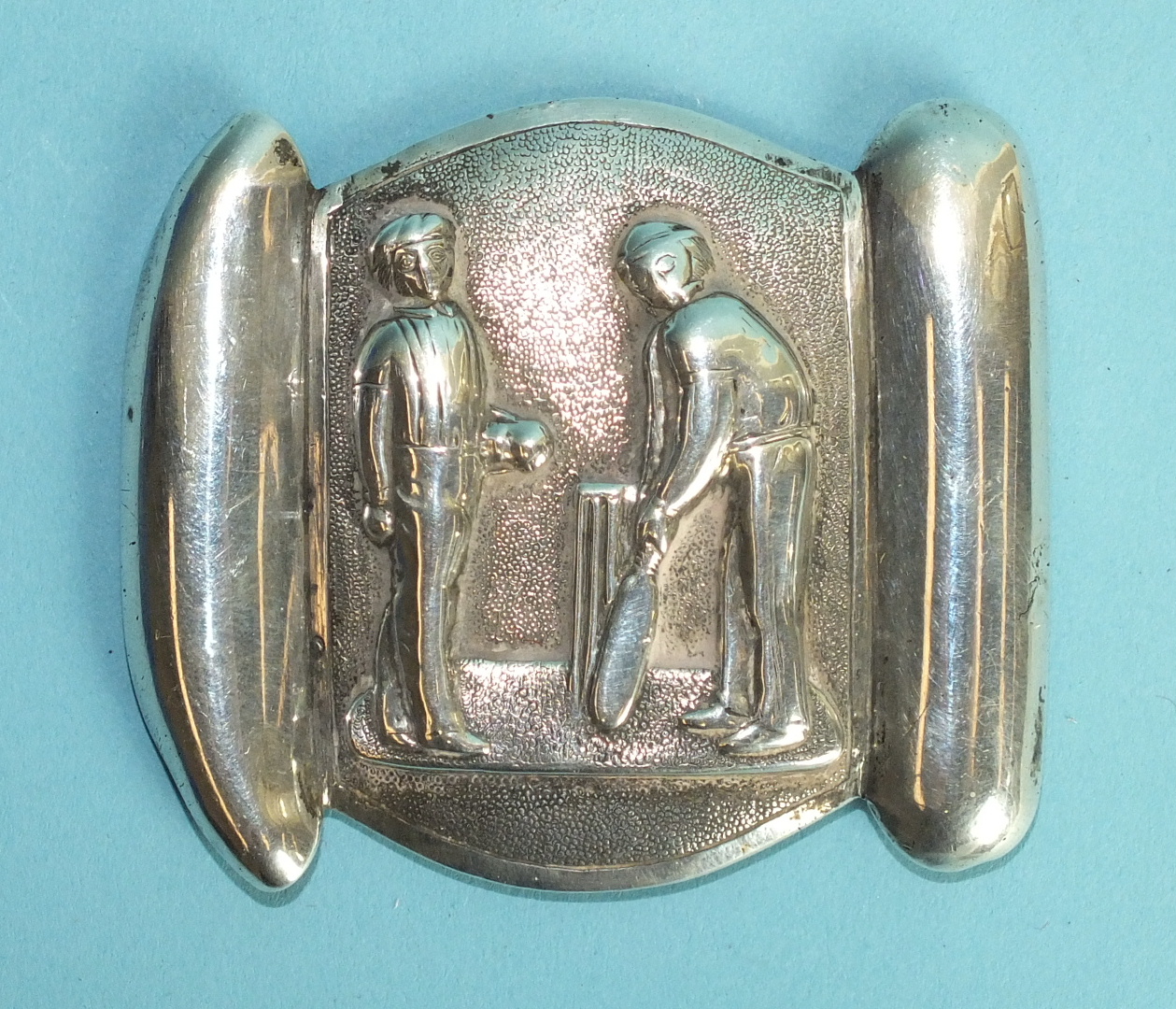 A white metal novelty belt clip cast as two cricketing figures, a bowler and a batsman before a