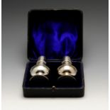 A cased pair of Edwardian silver pepper pots, each of plain bellied form to a filled and spread