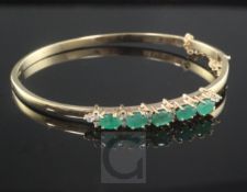 A 9ct gold, emerald and diamond hinged bangle, set with five oval cut emeralds.