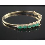 A 9ct gold, emerald and diamond hinged bangle, set with five oval cut emeralds.