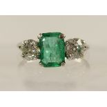 A platinum emerald and diamond ring the step cut emerald is claw set between two old cut diamonds of